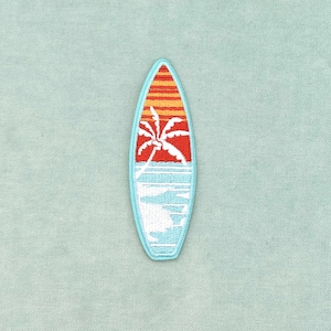 Peut inclure: Un écusson brodé en forme de planche de surf avec une bordure bleu clair. Le motif représente la silhouette d'un palmier blanc sur un ciel rouge et orange au coucher du soleil, au-dessus d'un océan bleu clair avec des vagues blanches.