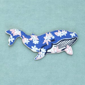 Peut inclure: Un écusson brodé en forme de baleine. La baleine est principalement bleue avec un ventre blanc orné de rayures verticales noires. Le corps est décoré d'un motif de petites fleurs blanches et rose clair sur toute la surface du design.
