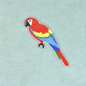 Peut inclure: Un écusson brodé thermocollant en forme de perroquet ara rouge. L'oiseau présente une tête et un corps rouge vif, des plumes d'ailes jaunes et vertes, ainsi qu'une longue queue bleue et rouge. Il possède une tache blanche sur le visage.