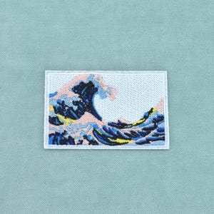 Puede incluir: Un parche rectangular bordado que presenta una representación estilizada de una gran ola oceánica. El diseño utiliza hilos de color azul oscuro, azul claro, amarillo y rosa pálido sobre un fondo blanco, creando una escena detallada de agua.