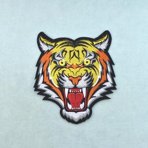 Op de afbeelding: Een geborduurde patch met de kop van een tijger die zijn bek open heeft in een brul. De tijger heeft gele vacht met zwarte strepen, oranje accenten, witte accenten en felgele ogen. Het ontwerp is omlijnd met een dikke zwarte rand.