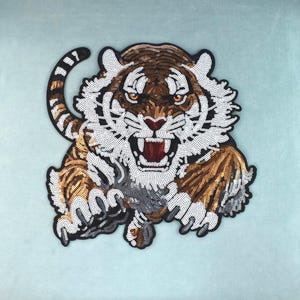 Può includere: Una toppa a forma di tigre realizzata con paillettes nei toni dell'arancione, nero e bianco. Il design raffigura una tigre ruggente con la bocca aperta che mostra denti rossi, strisce facciali dettagliate e una coda curva, bordata di nero.