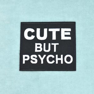 May include: A square black fabric patch with a stitched border featuring the white embroidered text CUTE BUT PSYCHO in a bold, sans-serif font. The patch is centred and displays clean, sharp lettering against the dark background.