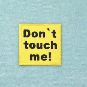 Pode incluir: Um emblema quadrado amarelo bordado com o texto preto Don't touch me! numa fonte sem serifa em negrito. O emblema tem uma borda costurada e é exibido sobre um fundo de tecido azul claro sólido com uma textura tecida visível.