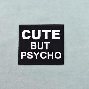 Peut inclure: Un écusson carré noir avec les mots "CUTE BUT PSYCHO" brodés en blanc. L'écusson est centré sur un fond de tissu bleu-vert clair. Le texte est en gras, en police sans empattement.