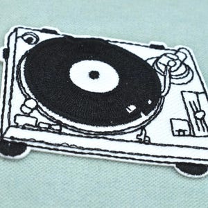 May include: A black and white embroidered patch featuring a detailed graphic of a turntable. The design includes a circular record on a rectangular base with visible tonearm, platter, and control sliders rendered in high-contrast thread work.