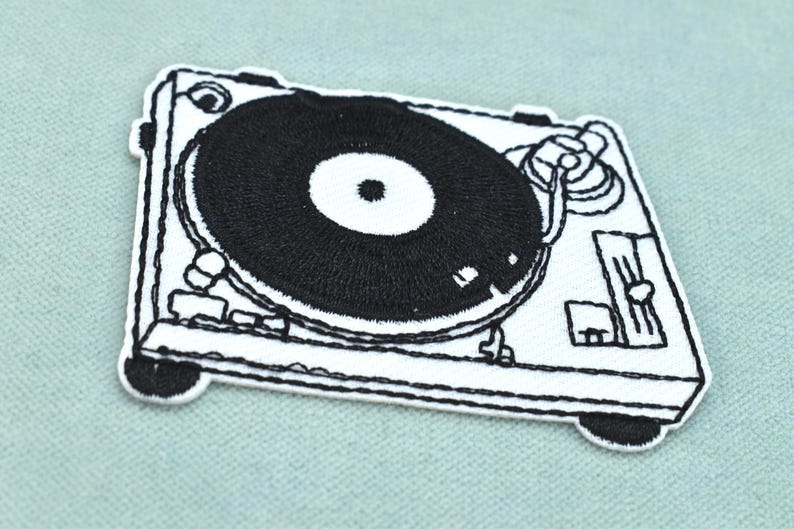 May include: A black and white embroidered patch featuring a detailed graphic of a turntable. The design includes a circular record on a rectangular base with visible tonearm, platter, and control sliders rendered in high-contrast thread work.