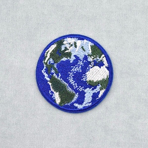 Könnte beinhalten: Ein runder, gestickter Aufnäher mit einer detaillierten Darstellung der Erde. Das Design zeigt Kontinente in Grün und Weiß vor einem blauen Ozean, mit einem dunkelblauen Rand. Geeignet für Kleidung oder Accessoires.