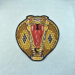 Può includere: Toppa ricamata raffigurante una testa di cobra con la bocca aperta, che mostra una lingua rossa e zanne. La testa del serpente è principalmente dorata e marrone, con dettagli neri e bianchi. Sfondo azzurro chiaro.