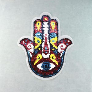 Peut inclure: Un écusson en forme de main de Hamsa orné de sequins aux couleurs vives, notamment rouge profond, jaune vif, blanc et bleu. Le centre de la paume présente un motif d'œil stylisé, le tout entouré d'une bordure en tissu blanc.