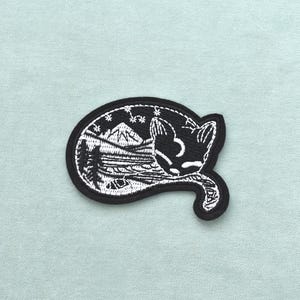 May include: Black and white embroidered patch depicting a sleeping cat with a mountain and starry night scene. The patch has a black border and is oval-shaped. The cat's tail curls around the design.