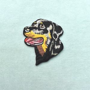 Peut inclure: Écusson brodé représentant la tête d'un chien Rottweiler. Le patch présente le chien avec des détails noirs, blancs, jaunes, marrons et roses. La langue du chien est visible, sur un fond bleu clair.