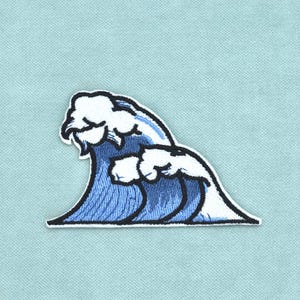May include: An embroidered patch featuring a stylised ocean wave design. The wave is rendered in shades of blue with white foam crests and a thick black outline, creating a graphic representation of a breaking wave on a light blue fabric background.