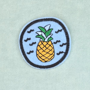 Op de afbeelding: Een ronde geborduurde patch met een gele ananas met groene bladeren, gecentreerd op een lichtblauwe achtergrond. Het ontwerp is omringd door kleine zwarte golfvormige symbolen en afgewerkt met een dikke, donkerbruin gestikte rand.