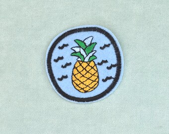 May include: A circular embroidered patch featuring a yellow pineapple with green leaves centered on a light blue background. The design is surrounded by small black wave-like symbols and is finished with a thick, dark brown stitched border edge.