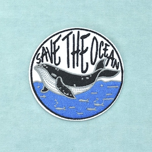 Pode incluir: Um emblema bordado circular apresentando uma baleia preta e branca nadando em água azul. O texto SAVE THE OCEAN está bordado em letras pretas acima da baleia. O emblema tem uma borda branca sobre um fundo azul claro.