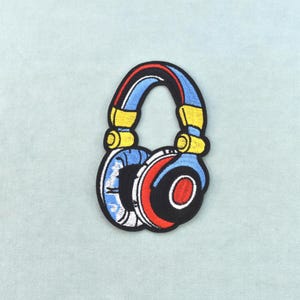 Op de afbeelding: Een geborduurde patch in de vorm van een koptelefoon. Het ontwerp bevat lichtblauw, geel, rood en zwart garen, met een dikke zwarte rand die de vorm van de koptelefoon omlijnt tegen een lichtblauwe stoffen achtergrond.