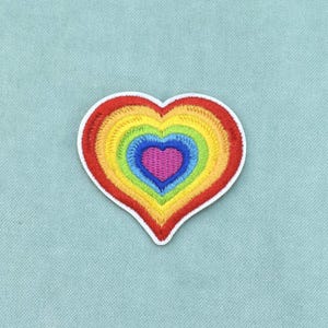 May include: An embroidered heart-shaped patch featuring concentric layers of color in a rainbow sequence. The center is a solid purple heart, surrounded by rings of blue, green, yellow, orange, and red, all finished with a white border edge.