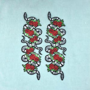 Può includere: Due toppe ricamate identiche che raffigurano un lungo serpente sinuoso a strisce bianche e nere. Il serpente è intrecciato con diverse rose rosse vivaci e piccole foglie verdi, creando un motivo verticale su ciascuna delle toppe.