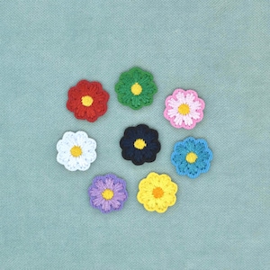 Pode incluir: Oito pequenas aplicações de flores de crochê circulares dispostas em círculo. As flores apresentam pétalas de várias cores, incluindo vermelho, verde, rosa, branco, preto, azul, roxo e amarelo, cada uma com um centro amarelo ou laranja contrastante.