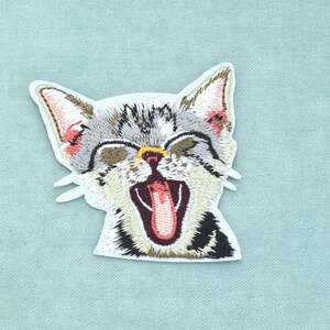 Op de afbeelding: Een geborduurde patch van een gapend kitten met grijze, witte en zwarte vacht. Het kitten heeft puntige oren, gesloten ogen en een open mond met een roze tong. De patch heeft een witte rand en is gevormd naar de kop van het kitten.