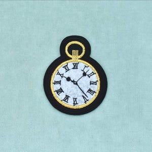 May include: An embroidered patch shaped like a pocket watch with a black border, a gold-coloured frame, and a white face. The watch face features black Roman numerals and two thin black hands pointing to the time, topped with a gold-coloured loop.