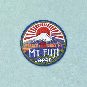 Könnte beinhalten: Ein kreisförmiger bestickter Aufnäher mit einer roten Sonne, die hinter dem schneebedeckten Berg Fuji aufgeht. Das Design zeigt ein rotes Torii, den Text 3776 m 12368 ft, MT FUJI und JAPAN in weißer und oranger Schrift auf blauem Rand.
