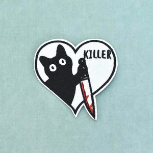 Può includere: Una toppa ricamata a forma di cuore che raffigura la sagoma di un gatto nero che tiene in mano un coltello argentato con macchie di sangue rosso sulla lama. La parola KILLER è ricamata in lettere maiuscole nere sopra il gatto su fondo bianco.