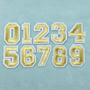 Op de afbeelding: Een set van tien opstrijkbare patches met de cijfers nul tot en met negen. Elk cijfer is uitgevoerd in metallic goudkleurig garen met een dikke witte rand, ontworpen in een klassiek varsity-bloklettertype met strakke randen en uniforme afmetingen.
