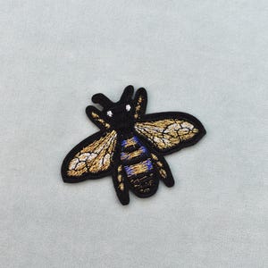 以下が含まれることがあります： 黒、金、青色の刺繍された蜂のパッチ。 蜂は詳細な羽と体を持っており、黒いアウトラインがあります。 パッチは水色の背景にあります。