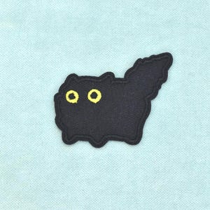 Peut inclure: Un écusson brodé noir en forme de chat touffu avec une grande queue dressée. Le chat possède deux yeux jaunes circulaires proéminents avec des pupilles noires, et le motif est entouré d'une bordure noire épaisse sur un tissu texturé.