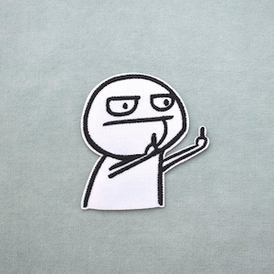 Puede incluir: Un parche bordado en blanco y negro que representa un personaje de meme con una expresión de enfado y el dedo corazón levantado. El parche tiene un borde negro y está sobre un fondo gris claro.
