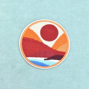 May include: A circular embroidered patch featuring a landscape design with a large dark red sun, layered orange and red hills, and a blue water element at the bottom. The patch has a thin orange border and is stitched with dense thread.