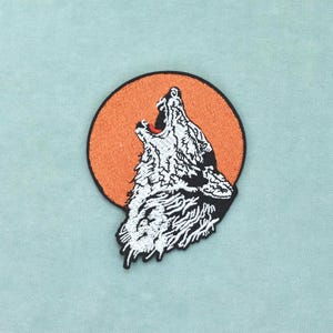 Peut inclure: Un écusson brodé circulaire représentant un loup blanc hurlant, la tête tournée vers le haut. Le loup est représenté en fil blanc avec des contours noirs sur un fond circulaire orange uni, le tout entouré d'une épaisse bordure noire.