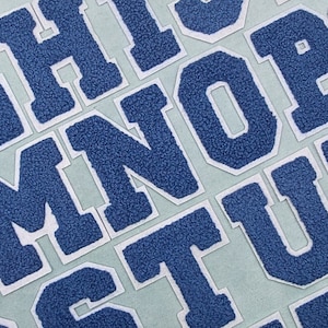 May include: Blue felt letters of the alphabet on a light blue background. The letters are in a variety of sizes and are arranged in a random order.