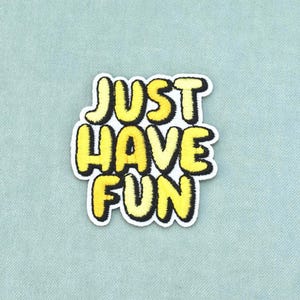 May include: An embroidered patch featuring the text JUST HAVE FUN in bold, yellow, block-style letters with a thick black outline. The lettering has a textured, fuzzy appearance and is set against a solid white background border.