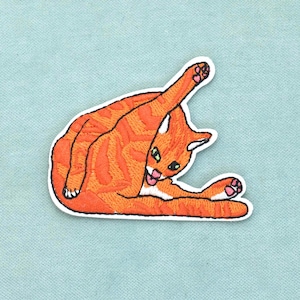 Op de afbeelding: Een geborduurde patch met een oranje cyperse kat met donkeroranje strepen, afgebeeld in een washouding met één poot omhoog. De kat heeft groene ogen, een klein roze tongetje en roze voetzooltjes, afgewerkt met een witte rand.