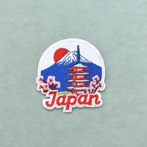 Peut inclure: Un écusson brodé circulaire représentant une montagne bleue au sommet enneigé blanc, un soleil rouge, une pagode rouge à plusieurs niveaux et des branches de cerisiers en fleurs roses. Le mot Japan est brodé en lettres cursives rouges.