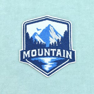Op de afbeelding: Een schildvormige geborduurde patch met een donkerblauw en wit berglandschap met een bos en een meer. Het woord MOUNTAIN is in witte hoofdletters geborduurd in het midden van de patch, die is voorzien van een witte rand.