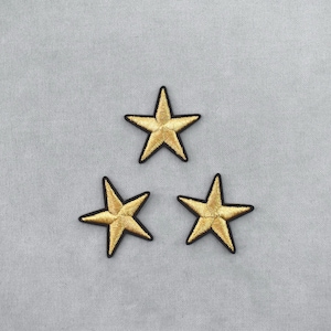 May include: Three gold star patches with black borders are arranged on a light gray fabric background. The stars have a metallic sheen, and the fabric has a subtle texture. These patches could be used for clothing or craft projects.