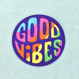 Peut inclure: Un écusson brodé circulaire avec un fond violet foncé présentant le texte GOOD VIBES en lettres grasses et épaisses. Les lettres sont remplies d'un motif multicolore de fils rose, orange, jaune, bleu clair et vert brodés.