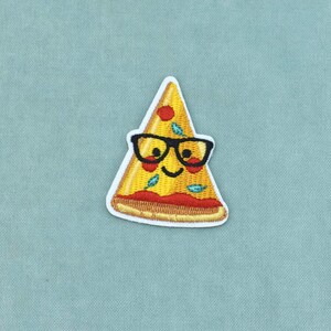 Op de afbeelding: Een geborduurde patch in de vorm van een pizzapunt met een gele korst, rode saus en groene toppings. De pizzapunt heeft een lachend gezicht en draagt een dikke bril met zwart montuur, alles omlijnd met een witte rand op een stoffen ondergrond.