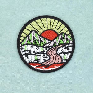 Peut inclure: Un écusson brodé circulaire représentant un paysage avec un soleil rouge, des rayons jaunes, des montagnes vertes et un chemin sinueux. Le motif inclut des détails blancs et noirs, le tout entouré d'une bordure circulaire noire épaisse.