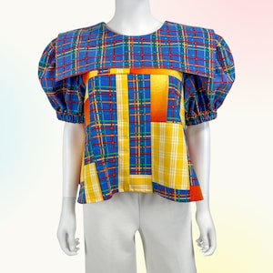 Puede incluir: Una blusa de cuadros azules y amarillos con un cuello grande y mangas abullonadas. La blusa tiene un patrón geométrico de rayas naranjas y amarillas.