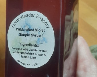 May include: A square bottle filled with a deep purple liquid, labeled "Wildcrafted Violet Simple Syrup" by Homesteader Soapery. The label lists ingredients: foraged wild violets, water, sugar, and lemon juice. The bottle contains 296 ml.