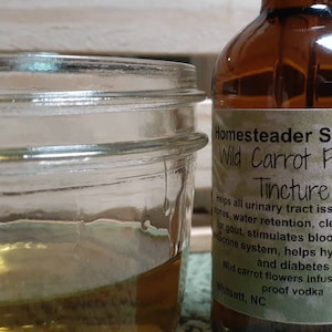 May include: A clear glass jar and a brown glass bottle with a label that reads "Homesteader Soapery Wild Carrot Flower Tincture." The jar contains a yellow liquid. The label lists health benefits.