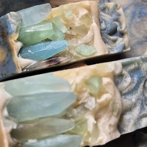 May include: Close-up of handmade soap bars with embedded blue and green crystal-like inclusions. The soap bars have a white base and blue and green swirls.