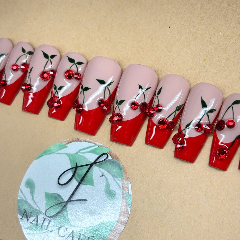 Peut inclure: Ensemble de dix faux ongles en forme de cercueil rouge et blanc avec un motif de cerises et des strass rouges.