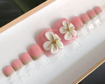 Könnte beinhalten: Ein Set künstlicher Zehennägel in einer Schachtel. Die Nägel sind rosa mit einer weißen Blume und einer goldenen Mittelverzierung. Die Nagelspitzen sind in einem gedämpften Grünton gehalten. Der Text "JSNAILCAFE" ist sichtbar.