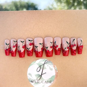 Peut inclure: Ensemble de dix faux ongles rouges et blancs avec un motif de cerises. Chaque ongle a une base rouge avec un bout blanc et une tige de cerise verte avec des cerises rouges. Les cerises sont d&eacute;cor&eacute;es de petits strass rouges.